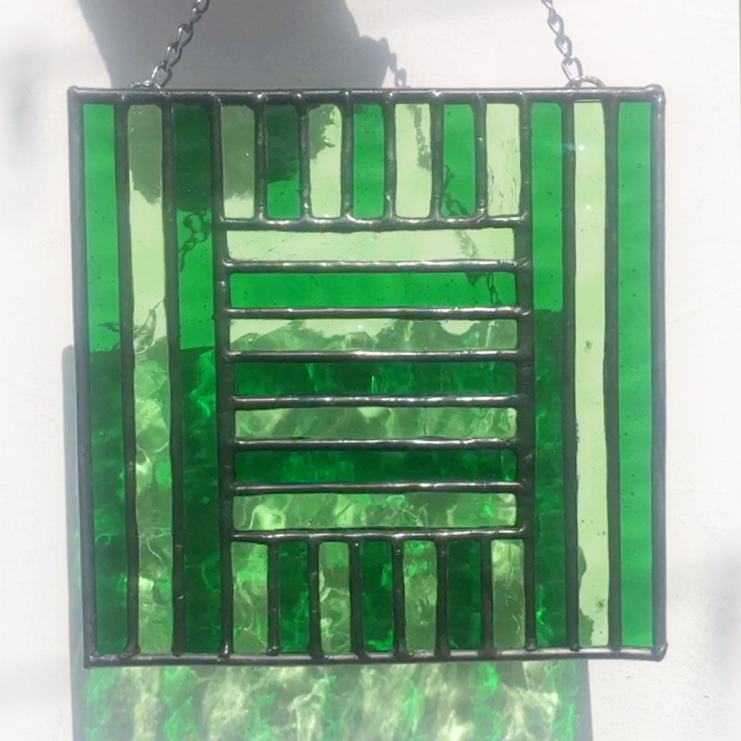 pale and deep green glass stripes are shaped to form a green glass square. Stained glass shown with sun shining through on white background. Green glass refractions are visible